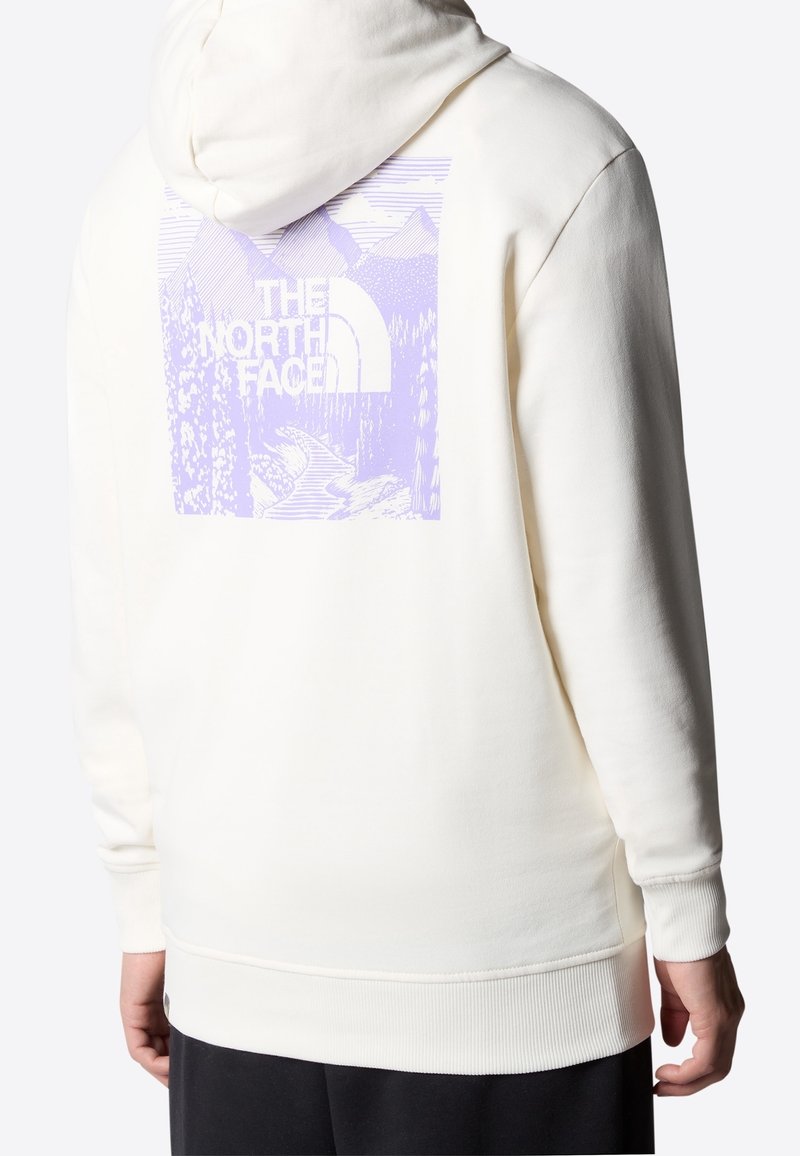 Personne portant un sweat à capuche blanc avec un graphisme de montagne violette, d'arbres, de rivière et le texte "THE NORTH FACE" dans le dos.