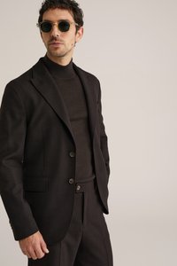 Homme portant un costume noir, un col roulé noir et des lunettes de soleil, debout devant un fond beige neutre.