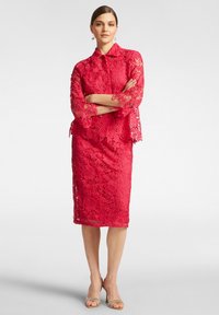 Abito rosso di pizzo con maniche a campana a 3/4 e gonna a matita. Motivi floreali intricati e bordi ondulati. Indossato con scarpe con cinturini color nude.