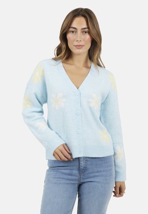 Donna che indossa un cardigan azzurro chiaro con motivi di margherite bianche e gialle, abbinato a jeans azzurri chiari, in piedi davanti a uno sfondo bianco.