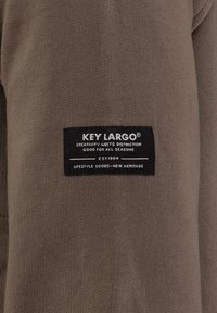 Textil marrón con una etiqueta rectangular negra y texto en blanco, que dice "KEY LARGO" y detalles de marca adicionales debajo.