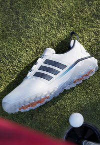 Chaussure de golf blanche avec des rayures noires sur gazon vert près d'une balle de golf positionnée au bord d'un trou sur le putting green.