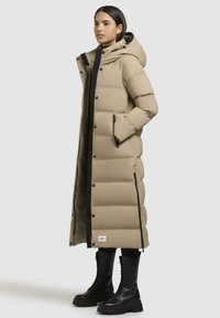 Beige lange gewatteerde jas met een capuchon, horizontaal gestikt patroon, zwarte accenten en ritszakken, gecombineerd met zwarte veterlaarzen.