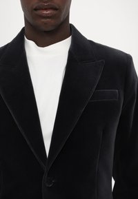 Jūrinės veliūro švarkas su įpjovimais apykaklėje, vienu mygtuko uždarymu ir krūtinės kišenė, dėvimas ant baltos marškinėlių. Sklandi tekstūra.