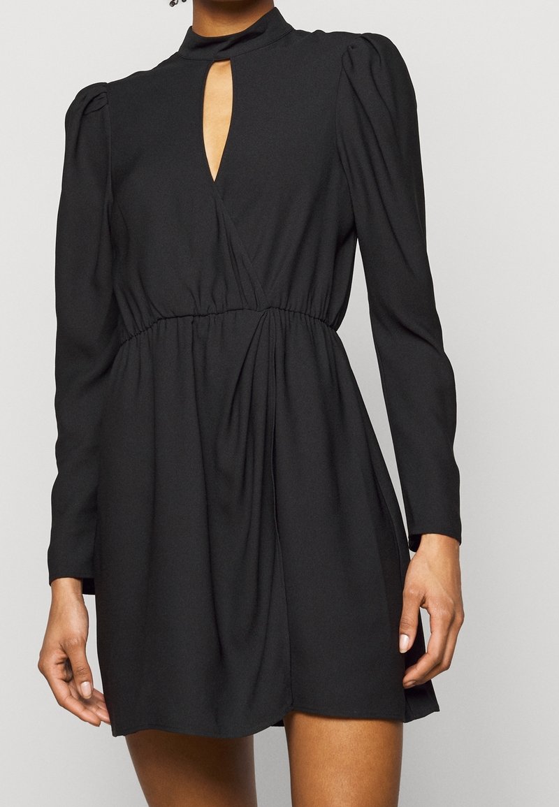Femme portant une robe noire à manches longues avec des épaules bouffantes, une taille cintrée et une encolure clé devant.