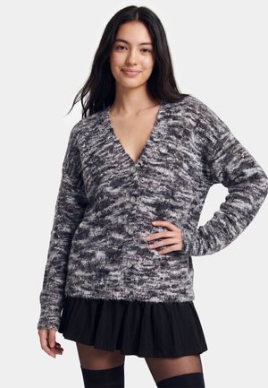 Jonge vrouw met lang donker haar die een grijs patroon gebreid vest met knopen en een zwarte rok draagt, staand met één hand op haar heup.