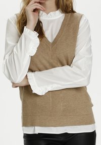 Hnědý pletený svetr přes bílou blůzu s krajkovým límcem a manžetami. Svetr má výstřih do V a jemný vzor rybí kosti.