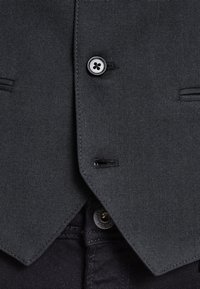 Chaleco gris oscuro hecho de tejido texturado, con un cierre de cuatro botones y detalles de costura sutiles, usado sobre pantalones negros.