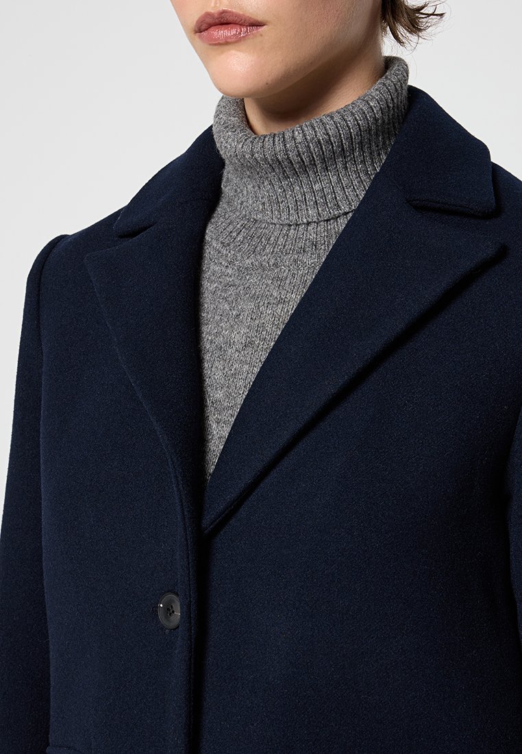 Marineblauer Wollmantel mit Reverskragen und einem einzigen Knopf, getragen über einem hellgrauen, gerippten Rollkragenpullover. Weiche Textur und klare Linien.