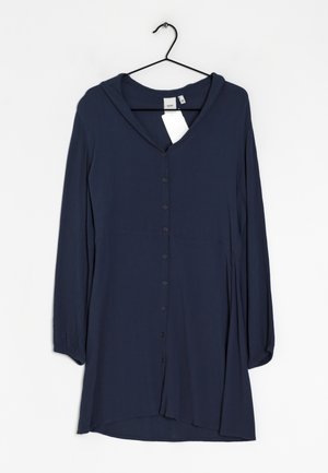 Robe chemise bleu marine à manches longues avec capuche, suspendue sur un cintre noir contre un fond blanc, avec des étiquettes de vêtement visibles.