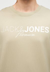 Μπεζ T-shirt με στρογγυλή λαιμόκοψη και λευκό λογότυπο "JACK & JONES Premium" στο στήθος, που φοριέται από ένα άτομο.