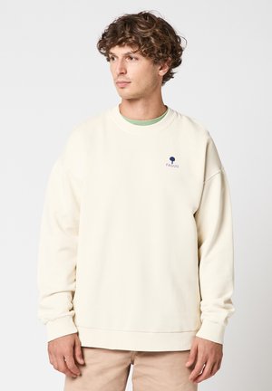 Jeune homme aux cheveux bouclés portant un sweat-shirt crème avec un petit logo d'arbre bleu et le texte « FAGUO » sur la poitrine.