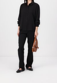 Black button-up shirt with long sleeves, paired with black trousers. Features flat black shoes with silver accents and a brown shoulder bag.