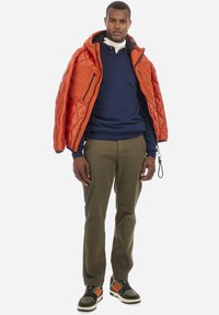 Puffjacka i ljus orange, quiltad design, med en marinblå sweatshirt under, olivgröna byxor och mönstrade sneakers.