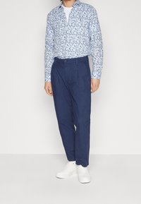 Světle modrá košile s květinovým vzorem, spojená s tmavě modrými kalhotami a bílými teniskami. Košile má dlouhé rukávy a límeček.