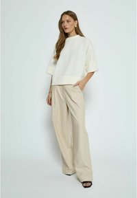 Biała, oversize bluza z krótkim rękawem zestawiona z beżowymi, szerokimi spodniami, które mają kieszenie i luźny krój. Czarne buty na odkrytej pięcie.