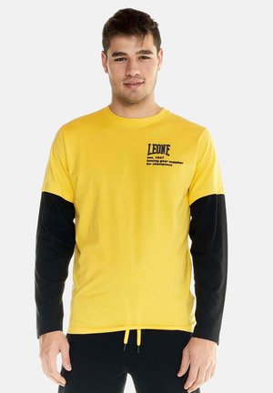 Junger Mann trägt ein gelb-schwarzes Langarmshirt mit dem Text "LEONE boxing gear supplier" auf der Brust, steht vor einem schlichten Hintergrund.