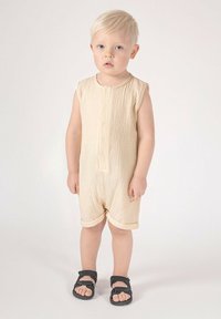 Beżowy kombinezon bez rękawów z zapinanym na guziki przodem i fakturowaną tkaniną. Szorty mają podwinięte mankiety. Na stopach czarne sandały z regulowanymi paskami.