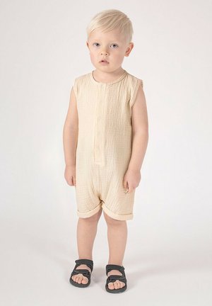 Beżowy kombinezon bez rękawów z zapinanym na guziki przodem i fakturowaną tkaniną. Szorty mają podwinięte mankiety. Na stopach czarne sandały z regulowanymi paskami.