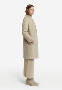 Cappotto in pelliccia sintetica beige con taglio dritto, chiusura frontale a bottoni e tasche laterali. Indossato sopra pantaloni a maglia beige e stivaletti con tacco.