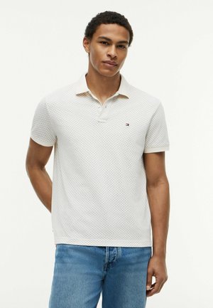 Jeune homme portant un polo blanc à motifs avec un petit logo et un jean bleu, debout devant un fond clair uni.