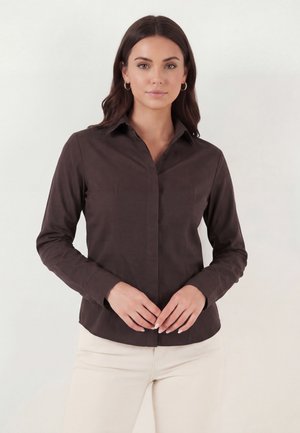 Kvinde med mørkebrunt hår iført små creoløreringe, en tætsiddende mørkebrun skjorte med knapper og lyse beige bukser, står med hænderne foldet.