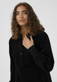 Camicia in velluto a coste nera con scollatura a placket, dettagli con bottoni e maniche lunghe con polsini aderenti. Tessuto morbido e testurizzato con vestibilità rilassata.