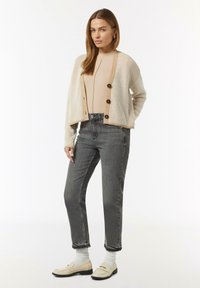Beige und cremefarbener gestreifter Cardigan mit dunklen Knöpfen über einem beigen Oberteil, kombiniert mit grauen Jeans und cremefarbenen Loafers. Weiche Texturen und taillierte Passform.