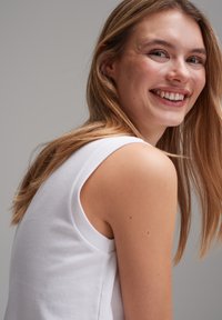White cotton sleeveless top with a round neckline, featuring smooth texture and subtle shoulder design. Hair is long and wavy.