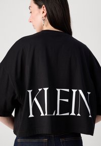 Černé zkrácené tričko vyrobené z bavlny s výrazným bílým nápisem "KLEIN" na zadní straně, s krátkými rukávy a volným střihem.