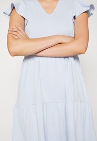 Vestido de algodão azul claro com decote em V e mangas curtas com babados, apresentando uma cintura franzida e uma saia rodada em camadas.