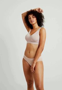 Egy göndör hajú nő világos bézs fehérneműben, az egyik karját felemelve pózol egy sima világos háttér előtt.