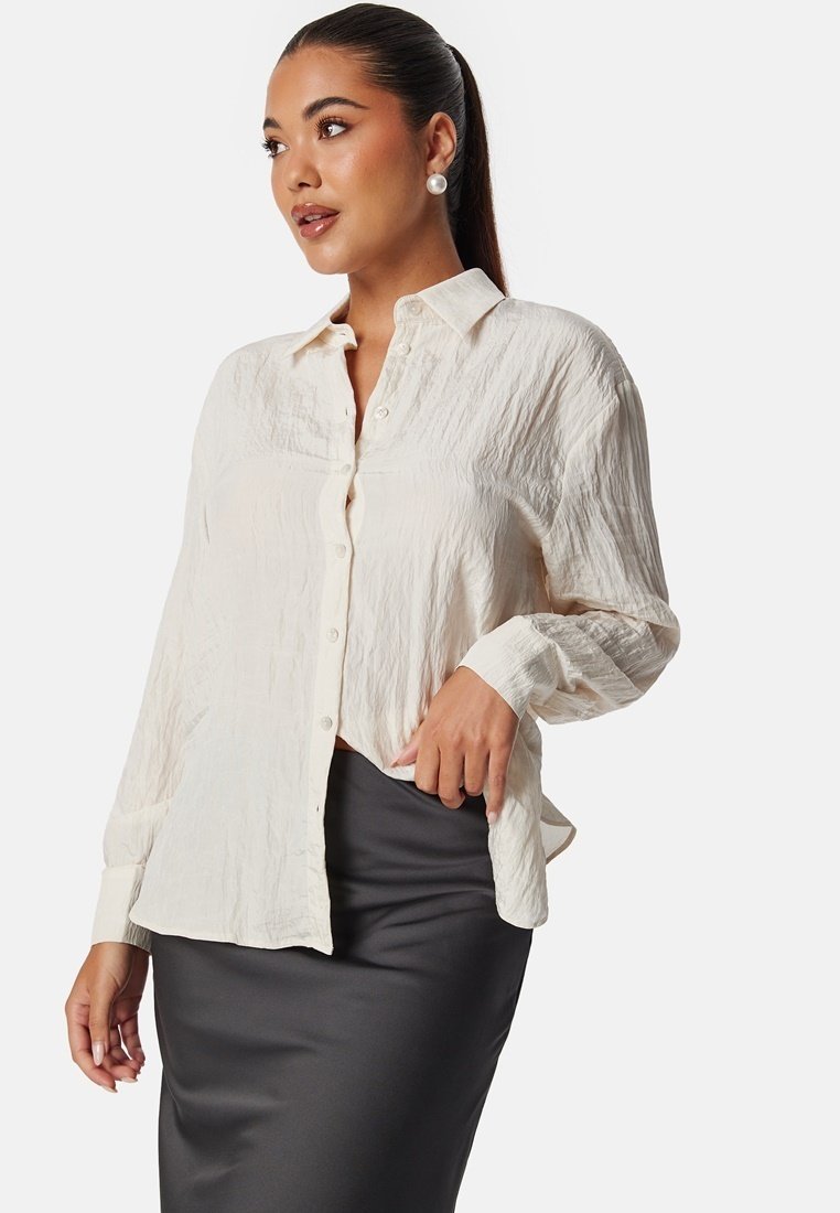Chemise boutonnée beige clair, froissée, avec un col et des manches longues, portée avec une jupe crayon gris foncé. Tissu texturé avec un léger éclat.