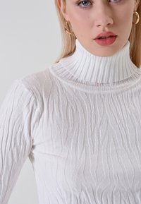 Primo piano di una persona che indossa un maglione a collo alto bianco e orecchini a cerchio d'oro, mostrando le labbra e parte del viso.