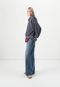 Overdimensioneret grå sweatshirt lagd over en rød ternet skjorte, kombineret med brede blå denim jeans og beige fodtøj. Modellen poserer sidelæns.