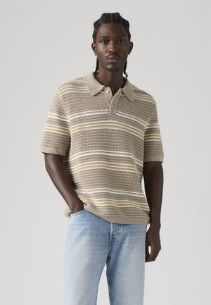 Hombre con cabello trenzado, vestido con una camiseta polo de punto a rayas beige y crema, y jeans azul claro, de pie con una mano en el bolsillo.