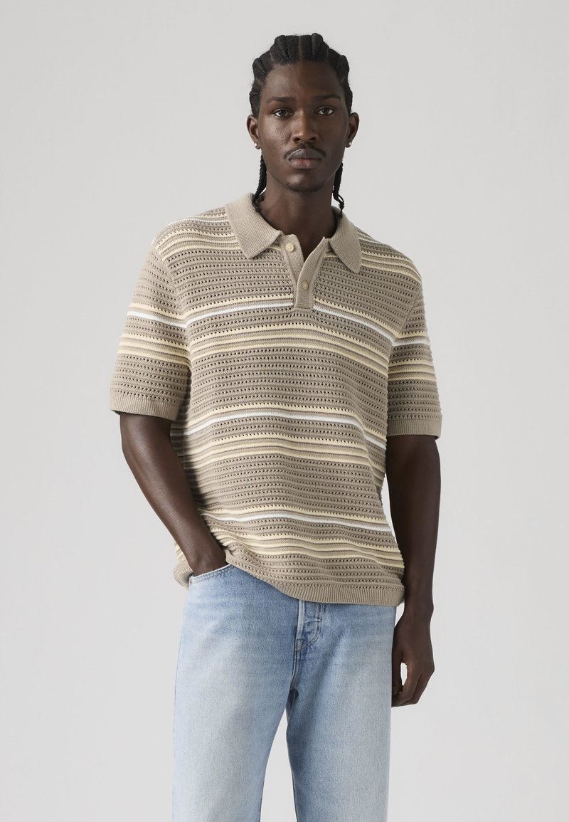 Homme aux cheveux tressés portant un polo en maille beige et crème à rayures, et un jean bleu clair, debout la main dans la poche.