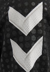 Svart strukturerad tyg med prickar har två överlappande vita zigzag-mönster med grå linjer. Designen skapar en lager-effekt.