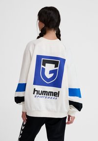 Vitt sweatshirt med en stor blå och svart grafik på baksidan, som har texten "hummel sportswear." Långa ärmar med blå och svarta ränder.