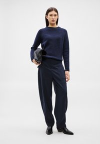 Γυναίκα που στέκεται μπροστά σε λευκό φόντο, φορώντας μπλε σκούρο πουλόβερ, φαρδιά μπλε σκούρα παντελόνια, μαύρα μποτάκια στον αστράγαλο, κρατώντας μια μαύρη διπλωμένη δερμάτινη ζακέτα.