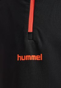 Schwarzer Sportstoff mit einer auffälligen roten Kordel und einem orangen "hummel"-Logo am unteren Rand. Glatte Textur, minimalistisches Design.