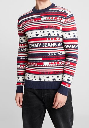 Man wearing a red, white, and blue patterned sweater with "TOMMY JEANS" text and star designs, paired with black pants.