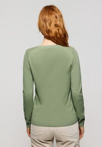 Pull vert à manches longues avec une texture lisse, des poignets et un ourlet côtelés, et un col rond, associé à un pantalon de couleur claire.