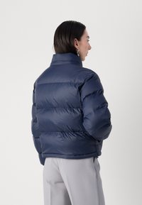 Navy blå dunjakke med høy krage og quiltet design. Glatt, blankt stoff med horisontal polstring og glidelås-lommer på sidene.