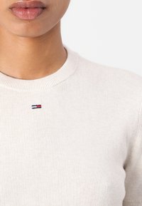 Svijetlosmeđi rebrasti pulover s okruglim izrezom. Ima mali izvezeni crveno-bijelo-plavi logo na prsima. Mekana tekstura.