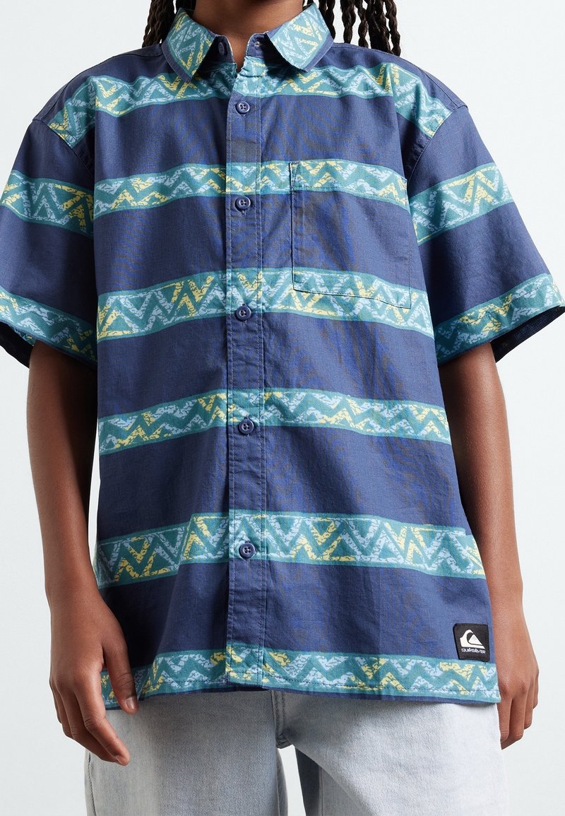 Personne portant une chemise bleue boutonnée avec des rayures en zigzag vertes et jaunes et un jean bleu clair.