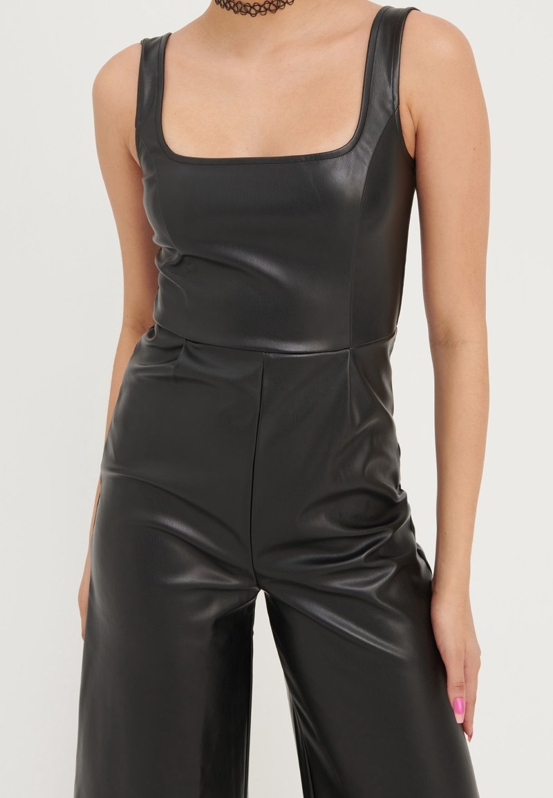 Svart jumpsuit i konstläder med fyrkantig halsringning, ärmlös design och lösa byxor. Slen yta med subtil lyster.