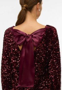 Bordeaux pailletten jurk met een diep decolleté op de rug, vastgebonden met een satin strik. De stof heeft een gestructureerd, glinsterend oppervlak.