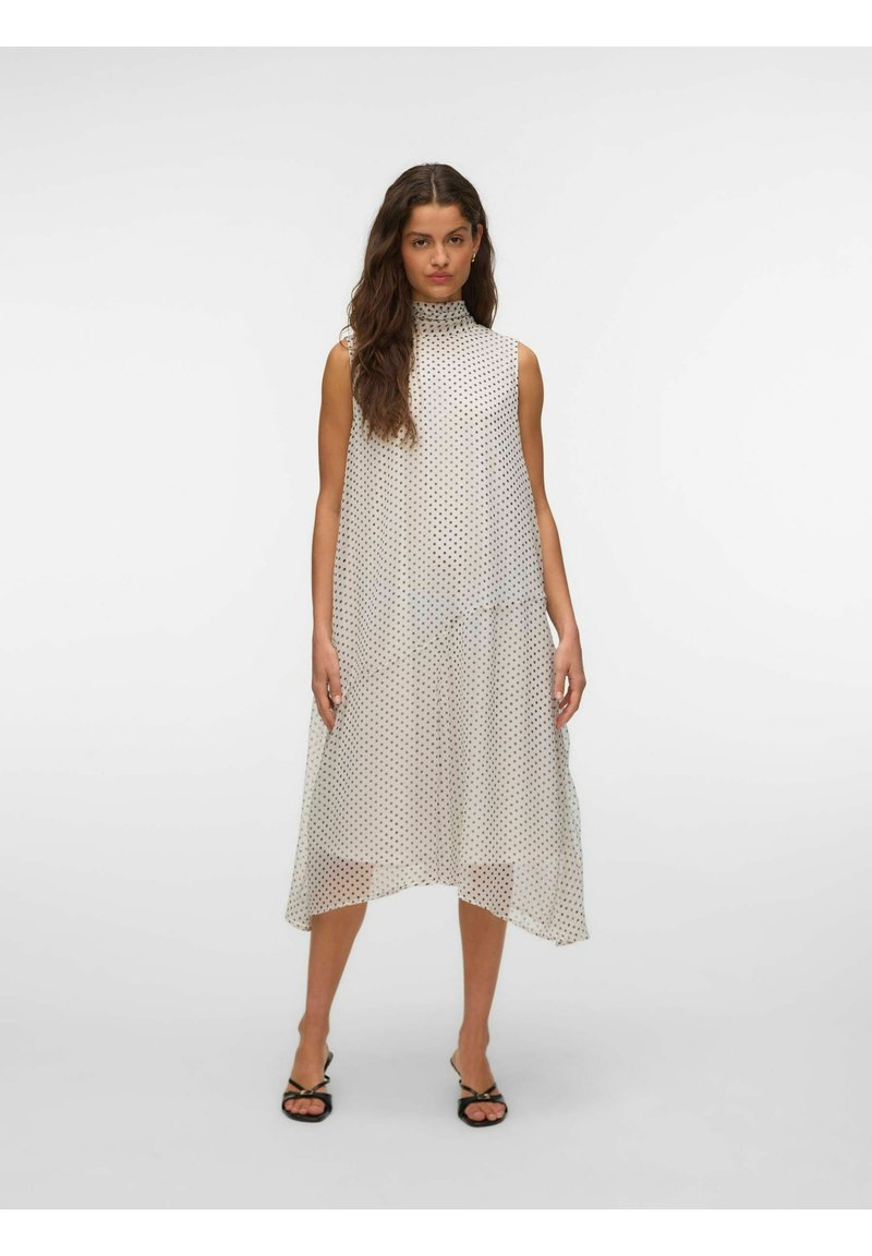 Femme aux longs cheveux foncés porte une robe midi blanche sans manches à petits pois noirs et des talons noirs à brides, debout devant un fond uni.