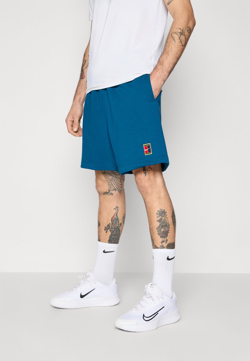 Calções de desporto turquesa com uma cintura elástica, bolsos laterais e um pequeno logótipo multicolorido. Usados com ténis e meias brancas da Nike.